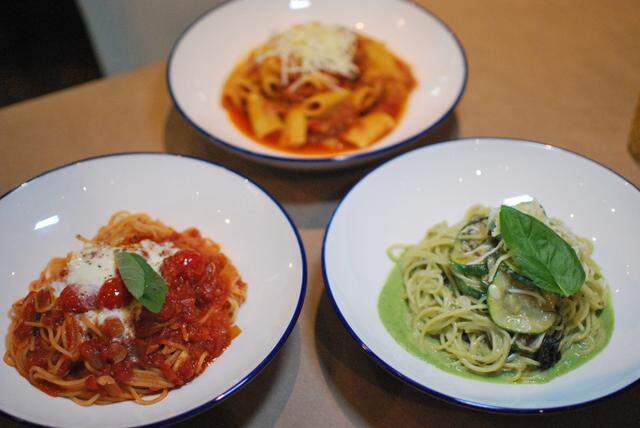A high-angle shot of three individual pasta dishes served in matching white bowls on a beige table. The dishes include a spaghetti dish with red tomato sauce and fresh mozzarella, a pasta dish with bright green sauce and vegetables, and a penne pasta dish topped with shredded cheese.