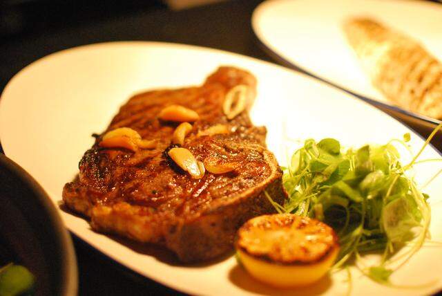 A close-up, warmly lit shot of a thick-cut, well-seared steak served on a white oval plate. The steak is topped with slices of roasted garlic and accompanied by a pile of fresh microgreens and a charred lemon half.