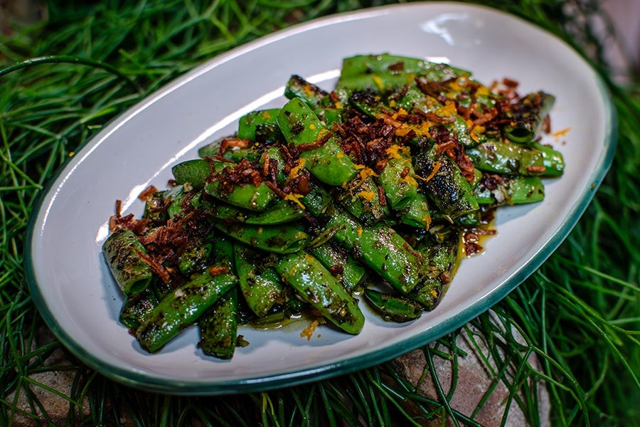 Grilled or sautéed green snap peas served on a white oval plate with a green rim, garnished with crispy fried shallots and a sprinkle of orange zest. The snap peas have char marks and a glossy coating, likely from oil or sauce.