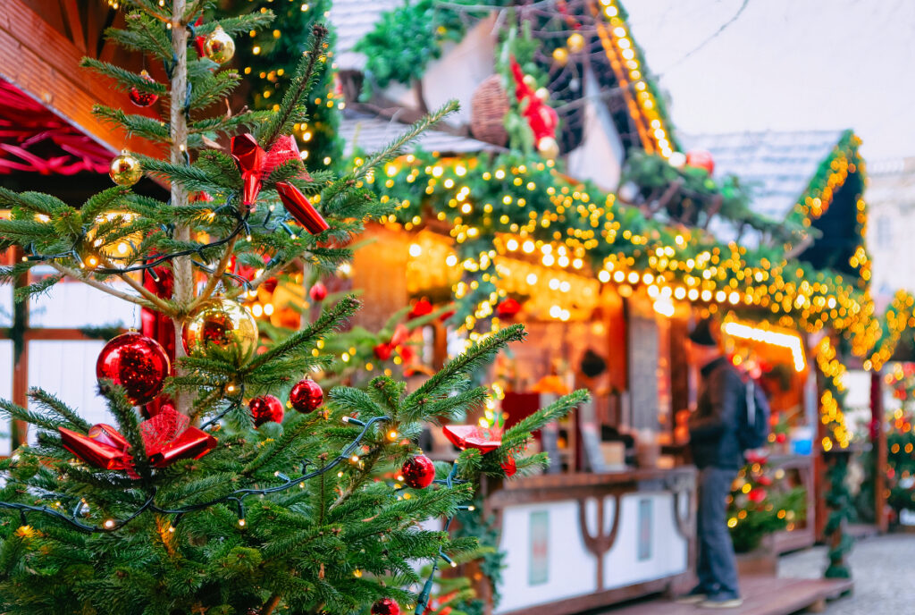 Image shows a Christmas tree with a decorated vendor in the background at a Christmas market.