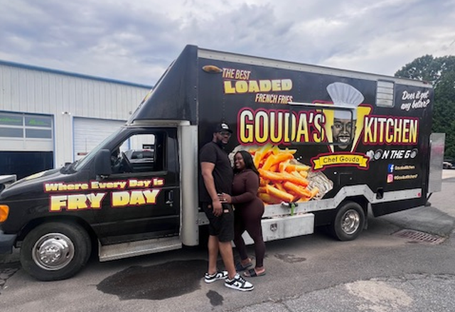 A man and a woman stand smiling together in front of a black food truck labeled “Gouda’s Kitchen: On The Go.” The truck’s vibrant wrap features a large image of loaded french fries and the playful slogan, “Where Every Day Is FRY DAY.” They are posed in a parking lot next to the vehicle, which also displays social media handles and an illustration of “Chef Gouda.”