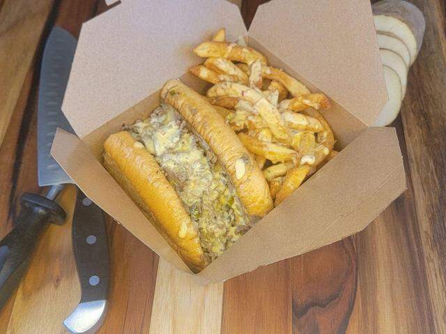 This takeout box contains a hearty meal featuring a large, stacked Philly cheesesteak sandwich overflowing with chopped steak, melted provolone, and sauteed peppers. Beside it are seasoned, crispy french fries lightly drizzled with the same creamy sauce, with a knife and sliced potato visible on the wooden board behind.