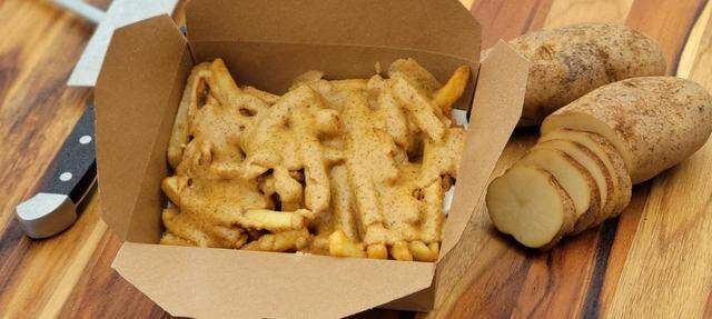 A brown cardboard takeout container is overflowing with golden, crispy french fries completely drenched in a thick, light-brown, peppered sauce, likely peanut sauce. The box rests on a wooden cutting board next to a chef’s knife, a whole Russet potato, and several freshly cut potato slices.