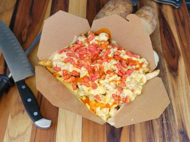 A unique and colorful dish of loaded fries is presented in a cardboard container, featuring fries smothered in a creamy white cheese sauce and a vibrant drizzle of bright red hot sauce. The entire creation is then generously topped with bright red, crumbled Flamin’ Hot Cheetos, positioned on a wooden surface next to raw potatoes and a knife.