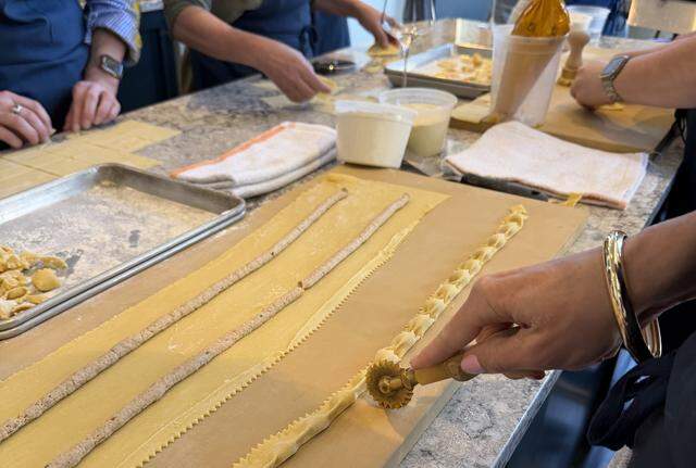 A close-up, high-angle shot of a communal pasta-making workshop or professional kitchen prep. Several people in blue aprons are gathered around a grey stone countertop, working on handmade stuffed pasta. In the foreground, a person’s hand uses a traditional brass fluted pasta wheel to cut along a long, folded sheet of golden egg dough. The dough has been filled with long, thin “logs” of a savory meat or vegetable filling, which are being pinched into individual pockets to create agnolotti.
