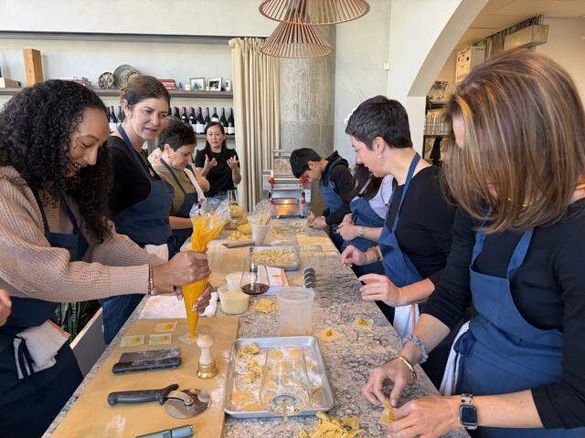 A wide-angle, eye-level shot of a lively, communal pasta-making workshop in a modern, sunlit kitchen. A group of diverse participants, all wearing navy blue aprons, are gathered around a long, grey-speckled stone countertop. In the foreground, a person with curly hair is carefully piping a vibrant orange squash filling from a large pastry bag onto small squares of fresh egg dough. To the right, other participants are actively folding the dough into stuffed shapes like tortelloni. The workspace is scattered with professional culinary tools, including a brass pasta stamp, a rolling cutter and trays of finished, hand-shaped pasta.