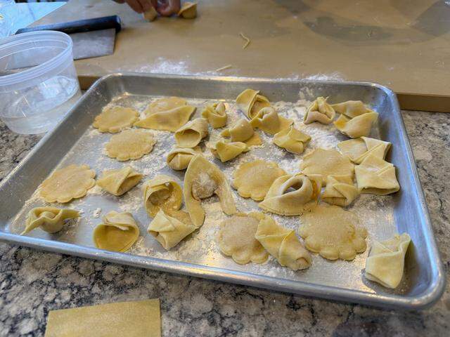 A close-up, high-angle shot focusing on a stainless steel sheet pan filled with various shapes of fresh, handmade stuffed pasta. The tray is lightly dusted with flour and sits on a grey-speckled stone countertop.