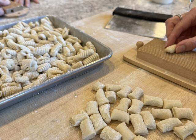 In the foreground, pillows of dough likely cut from a long “snake” sit waiting as a hand on the right is pressing one of those pillows against the rigagnocchi board.