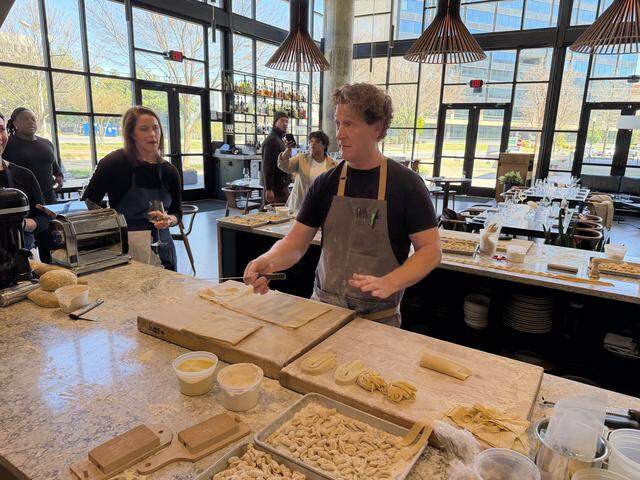 A wide, eye-level shot of a chef leading a pasta-making demonstration in a bright, modern restaurant. The chef with curly hair, wearing a grey apron, stands behind a large marble countertop covered in flour and various types of handmade pasta, holding a thin knife and gestuings toward sheets of fresh dough laid out on wooden cutting boards.
