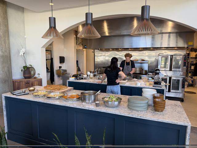 A wide shot of a modern, open-concept professional kitchen during a catered event or meal service. In the foreground, a large navy blue island with a grey speckled countertop holds several large stainless steel pans and pots filled with various pastas, a vibrant red tomato sauce, a hearty meat ragù, a large bowl of fresh green salad, and a thick, golden-brown slab of focaccia bread. To the right, stacks of grey and brown ceramic plates and bowls are ready for service.