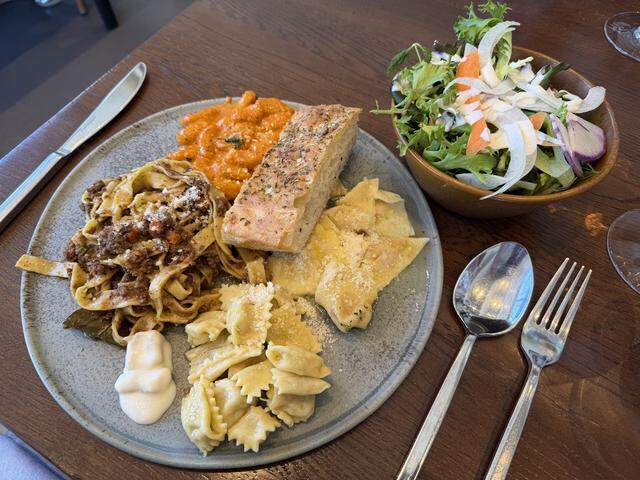 A high-angle, close-up shot of a full plate of handmade pasta and a side salad, representing the finished result of a pasta-making workshop. The grey ceramic plate features a sampler of several dishes: long ribbons of tagliatelle tossed in a dark, rich meat ragù; small agnolotti del plin in a light butter sauce; a portion of gnocchetti sardi in a vibrant orange-red sauce; and several large, folded cappellacci.