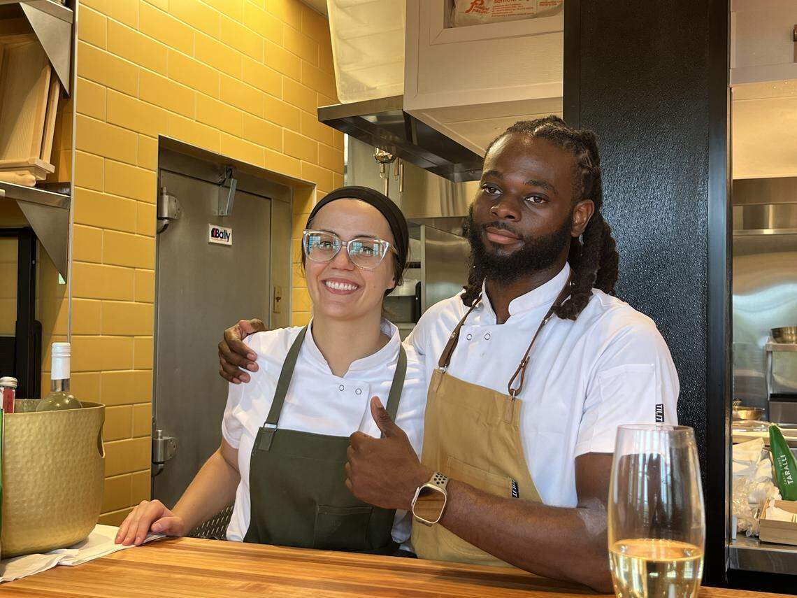 A portrait of a smiling couple standing behind a wooden counter in a professional kitchen. The person on the left, wearing glasses, a black head covering, and a green apron over a white chef’s jacket, is giving a thumbs-up. The one on the right, with dark hair in dreadlocks, is wearing a white chef’s jacket with a brown apron and has his arm around the other’s  shoulder. They are both looking directly at the camera. A glass of white wine is visible on the counter in the foreground. The kitchen has bright yellow tile walls and stainless steel appliances.