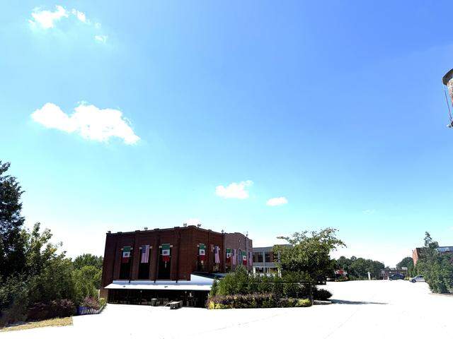 A long-distance, eye-level shot of a two-story brick building with flags hanging from the upper floor. The building is surrounded by a large, empty paved area under a vast blue sky with a few clouds.