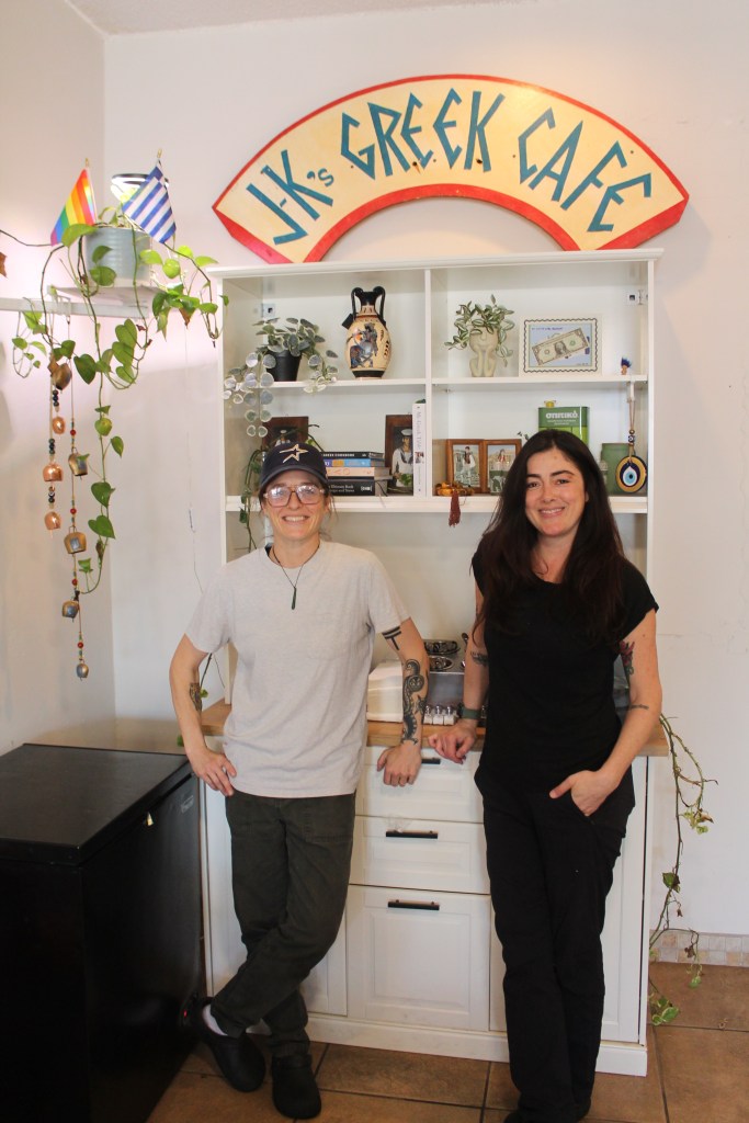 Two women pose next to shelves with plants and small items as well as a large J-K's Greek Cafe sign