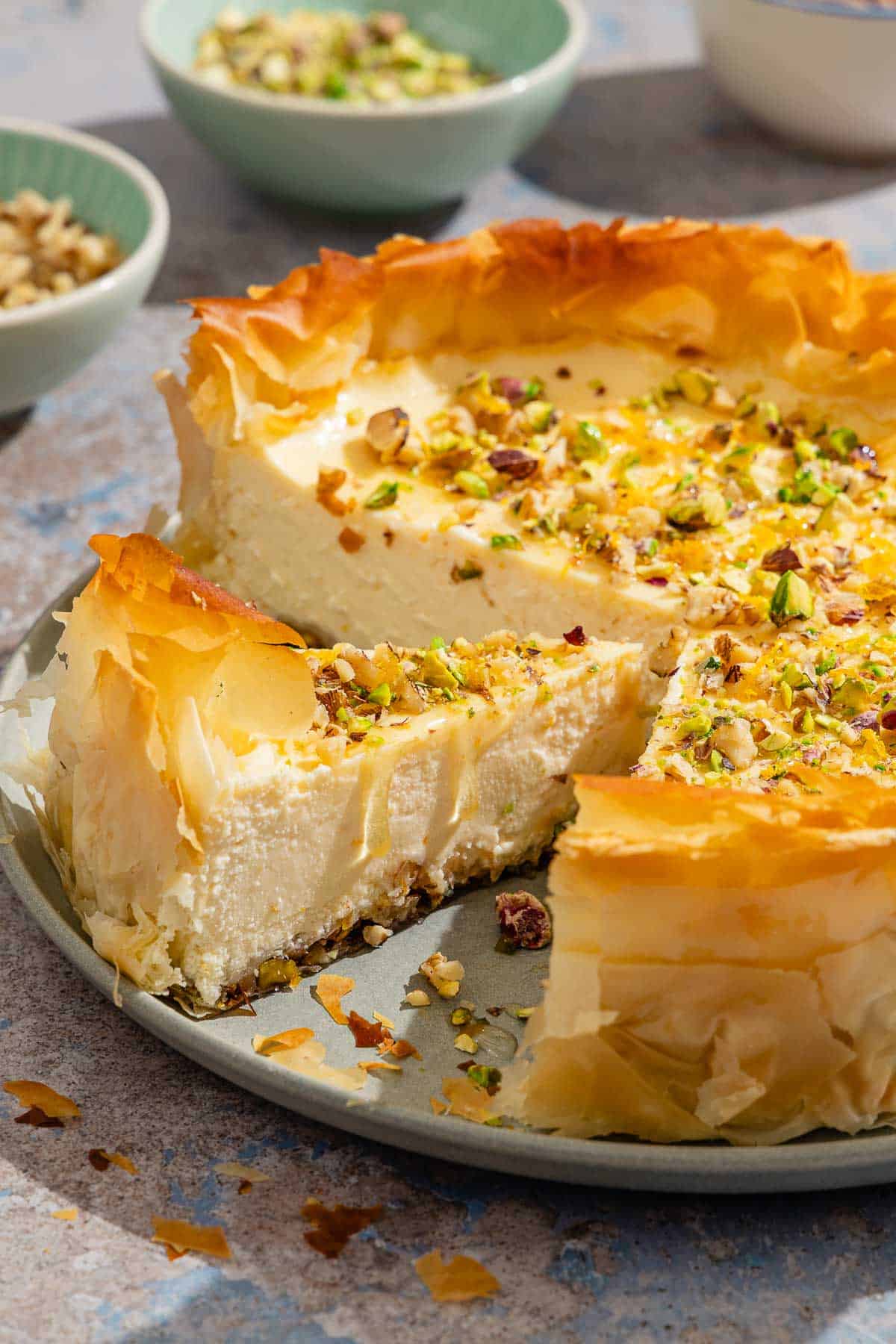 A close up of a slice of baklava cheesecake on a platter with the rest of the cake. I the background are small bowls of nuts.