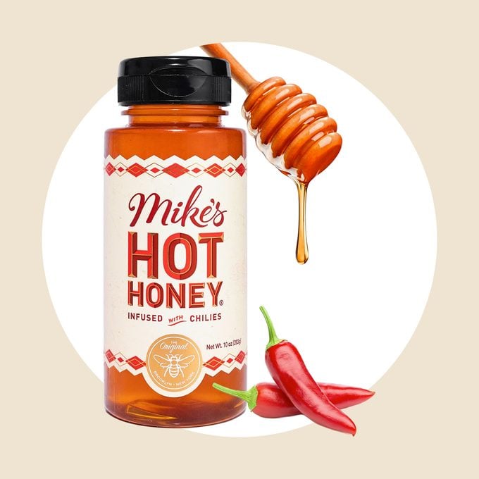 A bottle of Mike's Hot Honey is shown with a honey dipper dripping honey above it and two red chili peppers placed in front of the bottle, all on a light beige background.