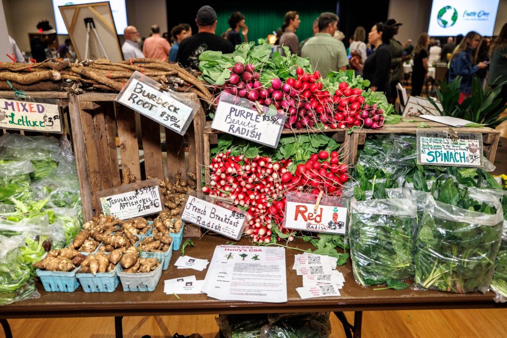 colorful piles of produce like radish and spinach are displayed in wooden crates with labels at an Earth Day farmers market at Illinois State University