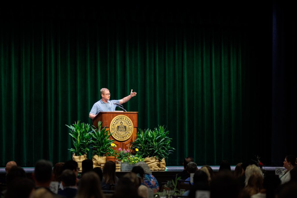 On a stage with vibrant green potted plants, Kelsey Timmerman speaks  and gestures at an Illinois State University podium during an Earth Day event