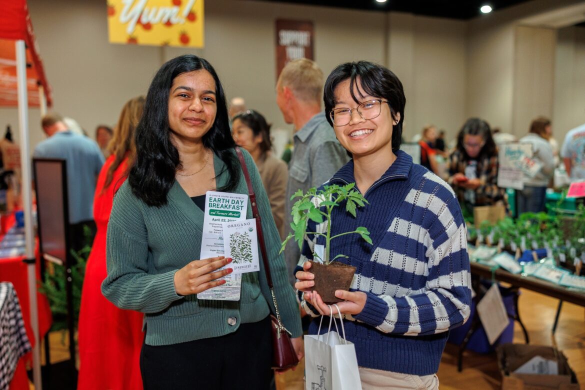 Two students smile at an Earth Day farmers market event at Illinois State University. One is holding fliers and the other is holding a plant