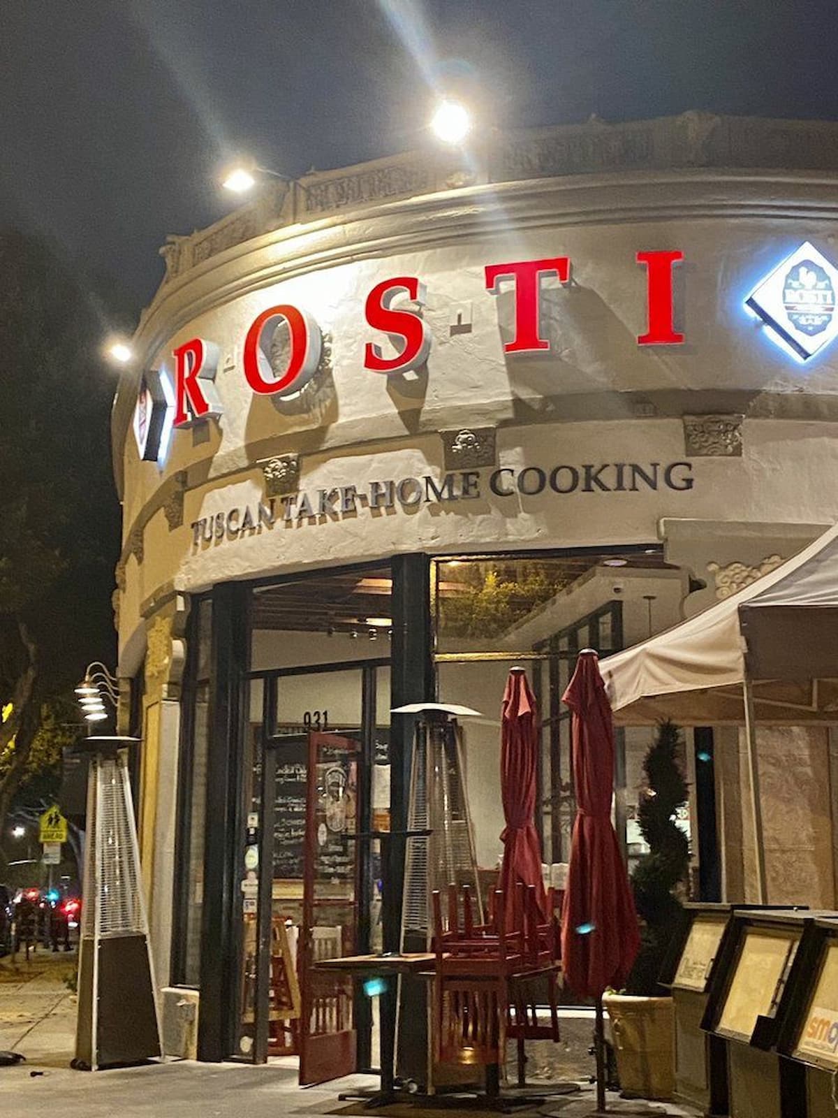 A restaurant exterior at night with bright red letters spelling "ROSTI," reminiscent of Italian chain restaurants in America, and a sign below reading "Tuscan Take-Home Cooking." Outdoor heaters and closed red umbrellas are set up near the entrance.