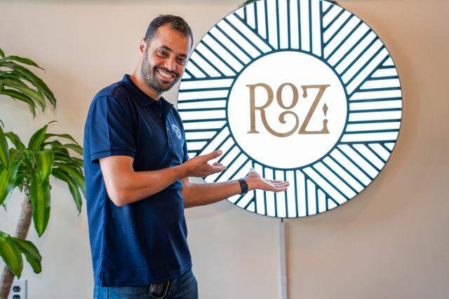 A man in a navy blue polo shirt stands smiling and gesturing with both hands toward a large, circular backlit sign on the wall. The glowing white sign features the word “ROZ” in gold lettering, surrounded by a complex geometric pattern of black radiating lines.