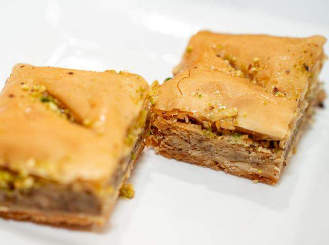Two square pieces of golden-brown baklava are presented on a white plate, showing their many thin, flaky layers of phyllo pastry and a thick nut filling. Each piece is lightly drizzled with honey syrup and garnished with a sprinkling of finely crushed green pistachios.