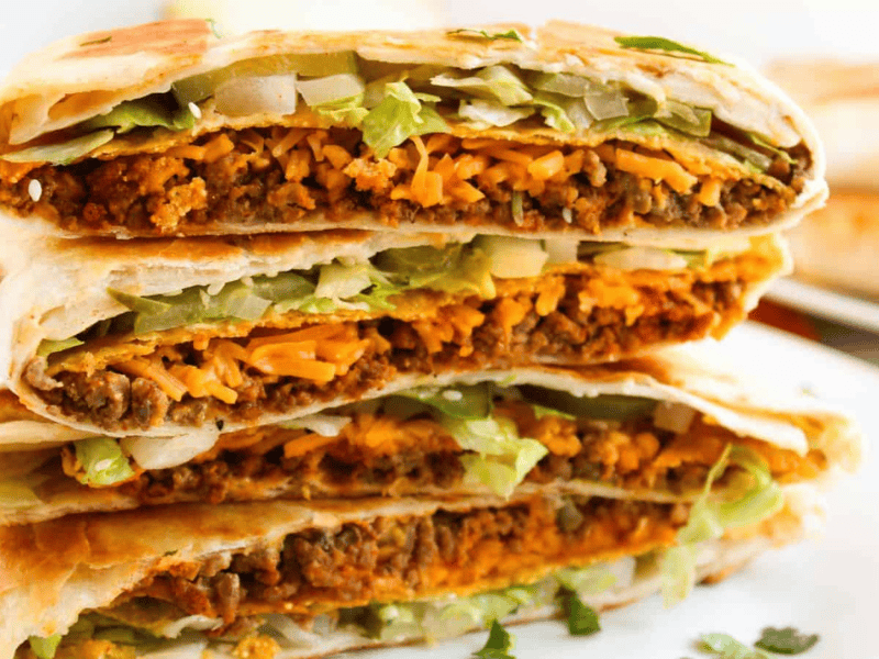 Close-up of a stack of folded tortillas filled with beef, melted cheese, lettuce, and onions on a white plate.