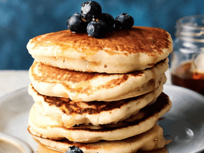 A stack of pancakes topped with blueberries and syrup sits on a plate, with a jar of syrup in the background.