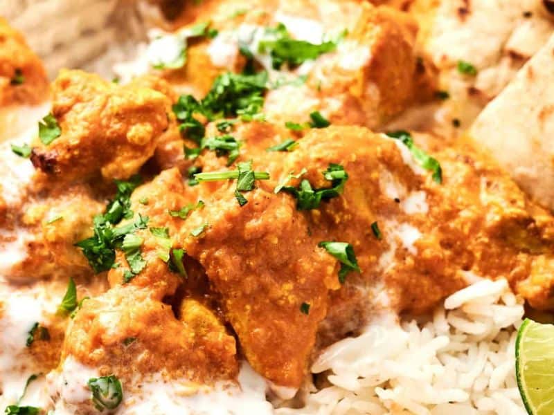 A close-up of chicken tikka masala served on rice, garnished with fresh cilantro, with lime wedges and naan on the side.