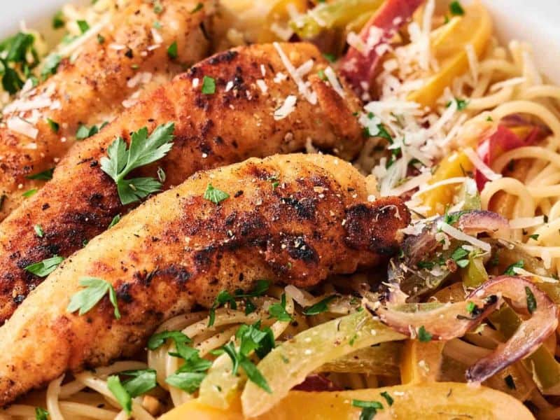 A plate of spaghetti topped with grilled chicken strips and garnished with chopped parsley, bell peppers, and shredded cheese.