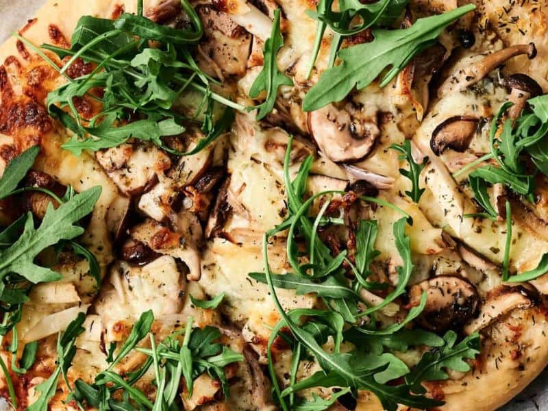 Close-up of a pizza topped with arugula, mushrooms, cheese, and herbs on a parchment paper.