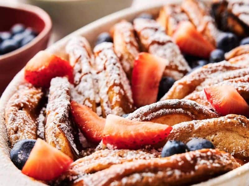 A baked French toast casserole topped with powdered sugar, sliced strawberries, and blueberries in a white dish.
