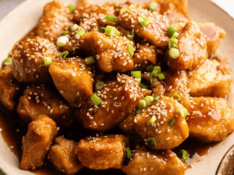 A plate of sesame chicken pieces covered in sauce, garnished with sesame seeds and chopped green onions.