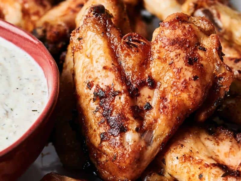 Close-up of grilled chicken wings with a crispy texture, next to a red bowl of creamy dipping sauce.