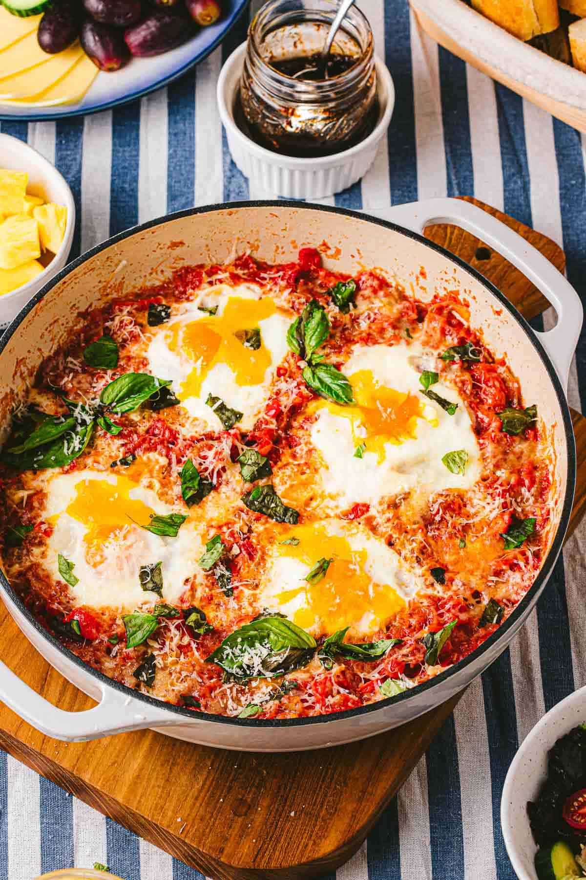 A skillet of eggs in purgatory next to a jar of jam and other various table settings.