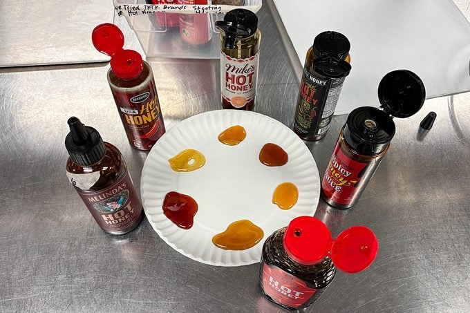 A white paper plate with seven different hot sauces or seasonings in small puddles, surrounded by their bottles on a stainless steel table. Each sauce varies in color from red to amber.