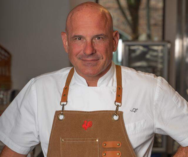 A close-up portrait of a bald chef wearing a white chef’s coat and a brown apron with leather straps and a red rooster logo. The chef is smiling and looking directly at the camera in a professional kitchen setting.