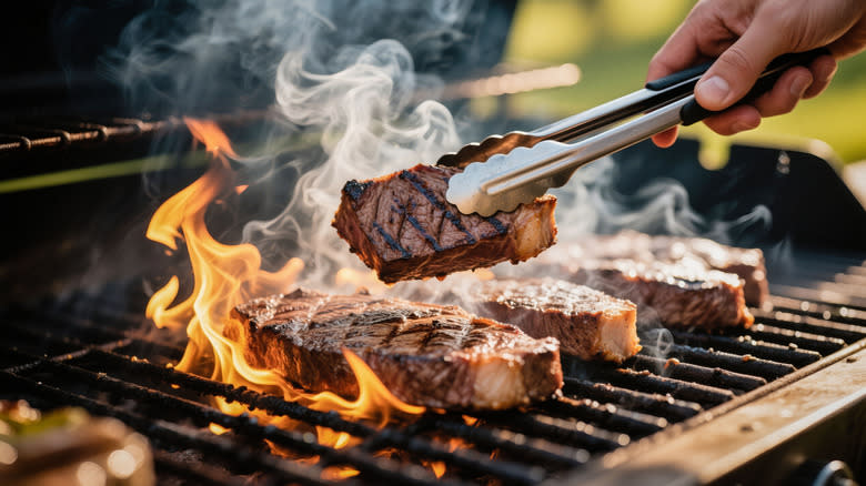 A hand holding tongs while flipping steak on the grill.