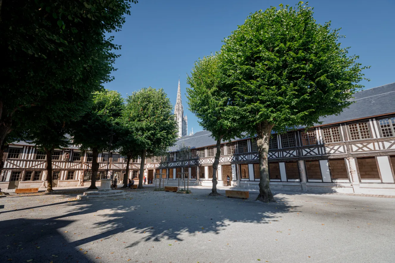 Aitre Saint-Maclou, a historical building in Rouen, France, featuring a courtyard with trees and a prominent steeple in the background.