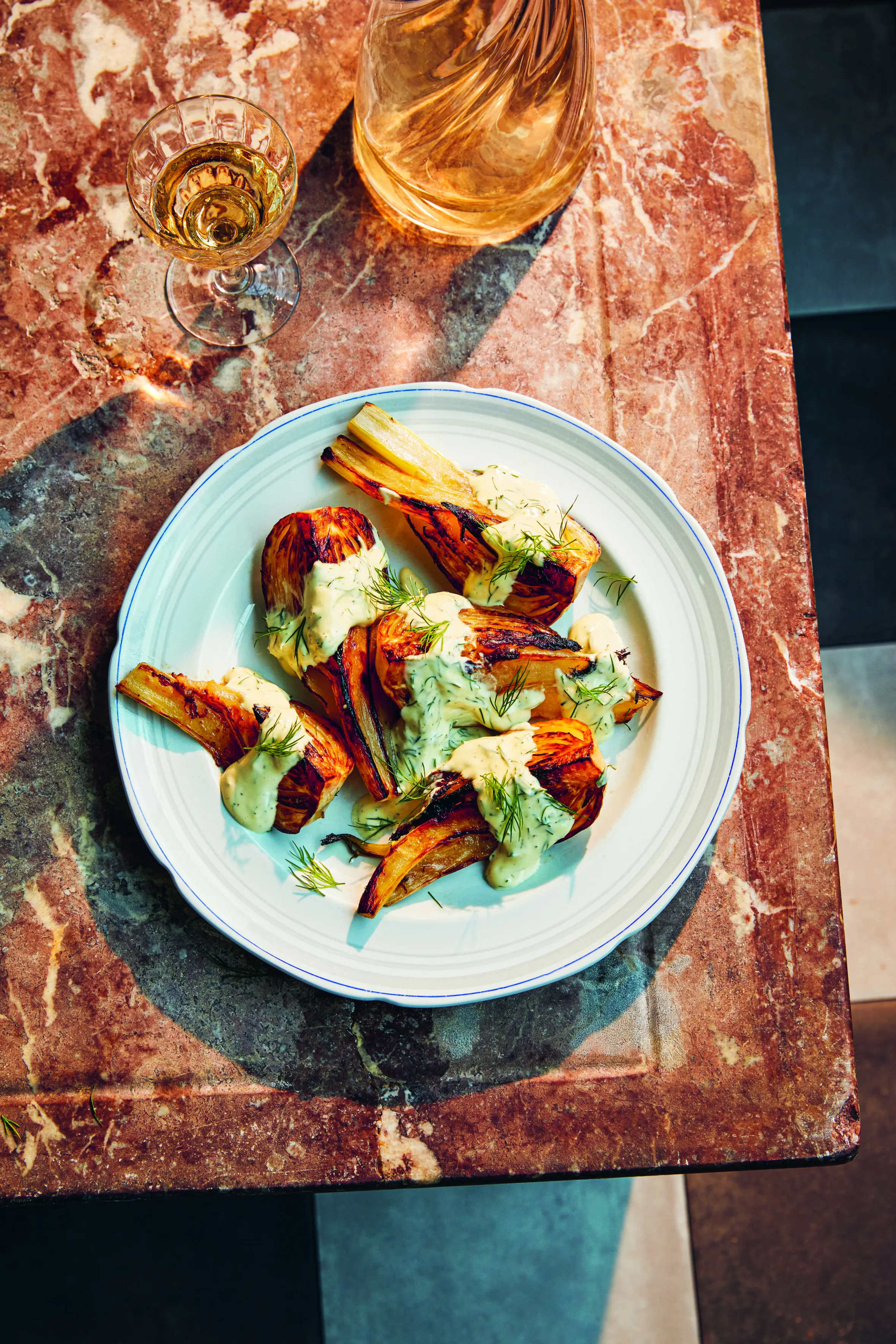 Fennel a la Grecque on a white plate with a glass of wine and a decanter on a marble surface.