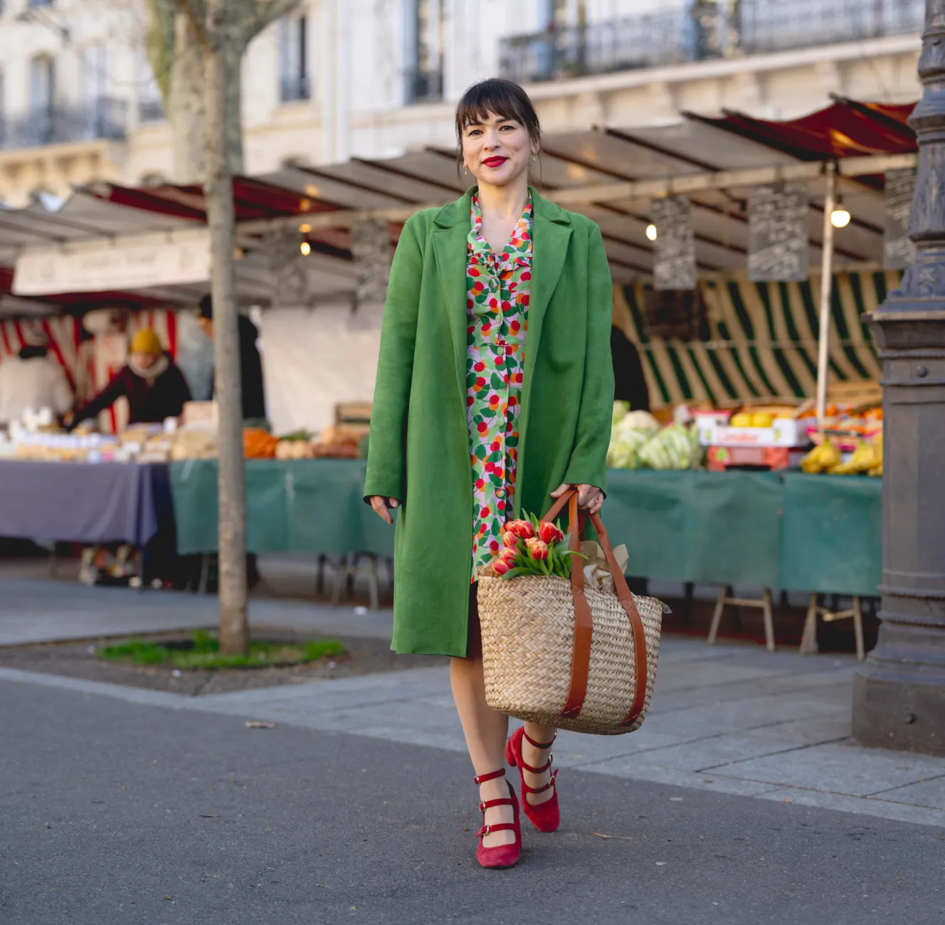 Rachel Khoo in a green coat and red shoes, holding a woven bag with tulips, walking past market stalls.