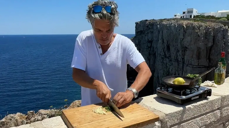Peter Hanley of Pete's Pans dices vegetables next to a pan on a portable stove, with the ocean and a cliff-face in the background