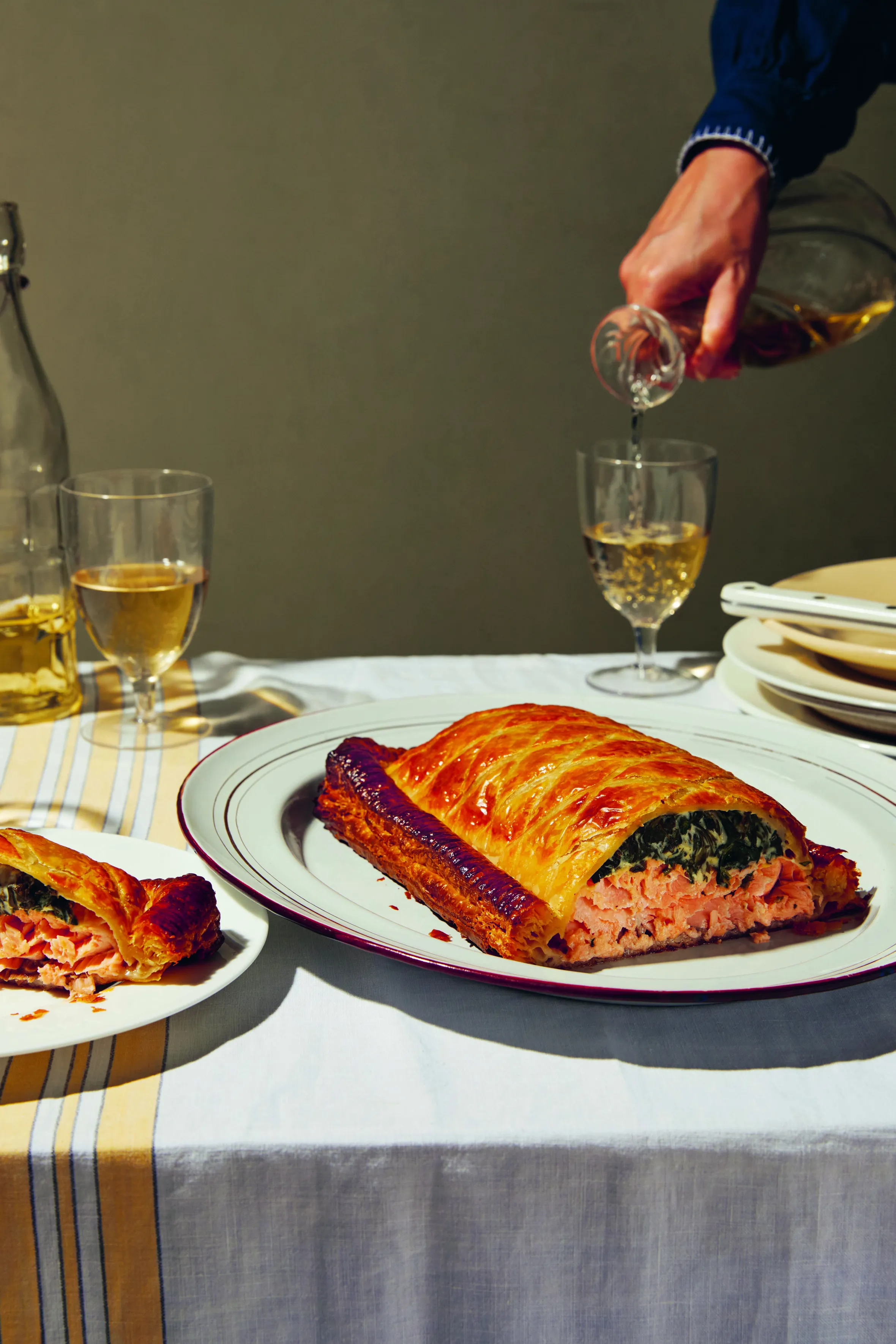Salmon en croute with spinach, served on a white plate next to a slice on a smaller plate, with a hand pouring wine into a glass.