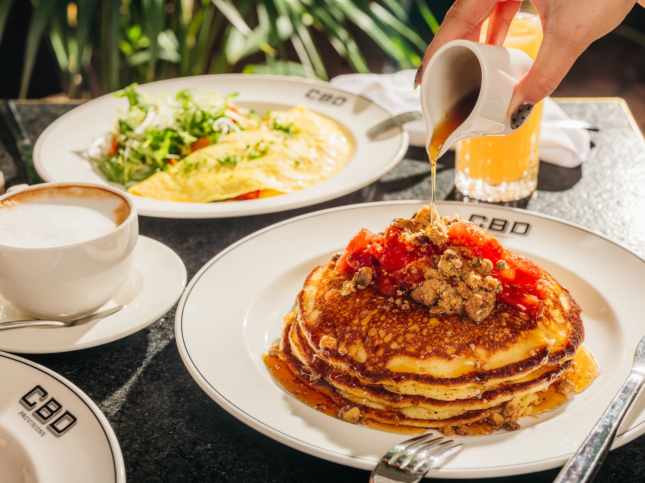 Stack of pancakes with syrup and topping, omelette with salad, cappuccino, and orange juice on table.