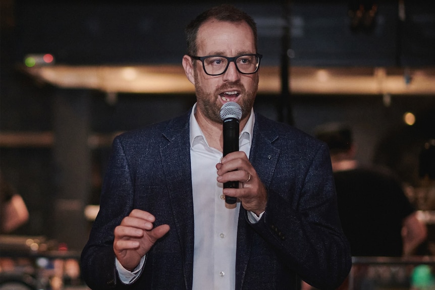 A bearded man in glasses and a suit speaks into a microphone while standing in what appears to be a pub.