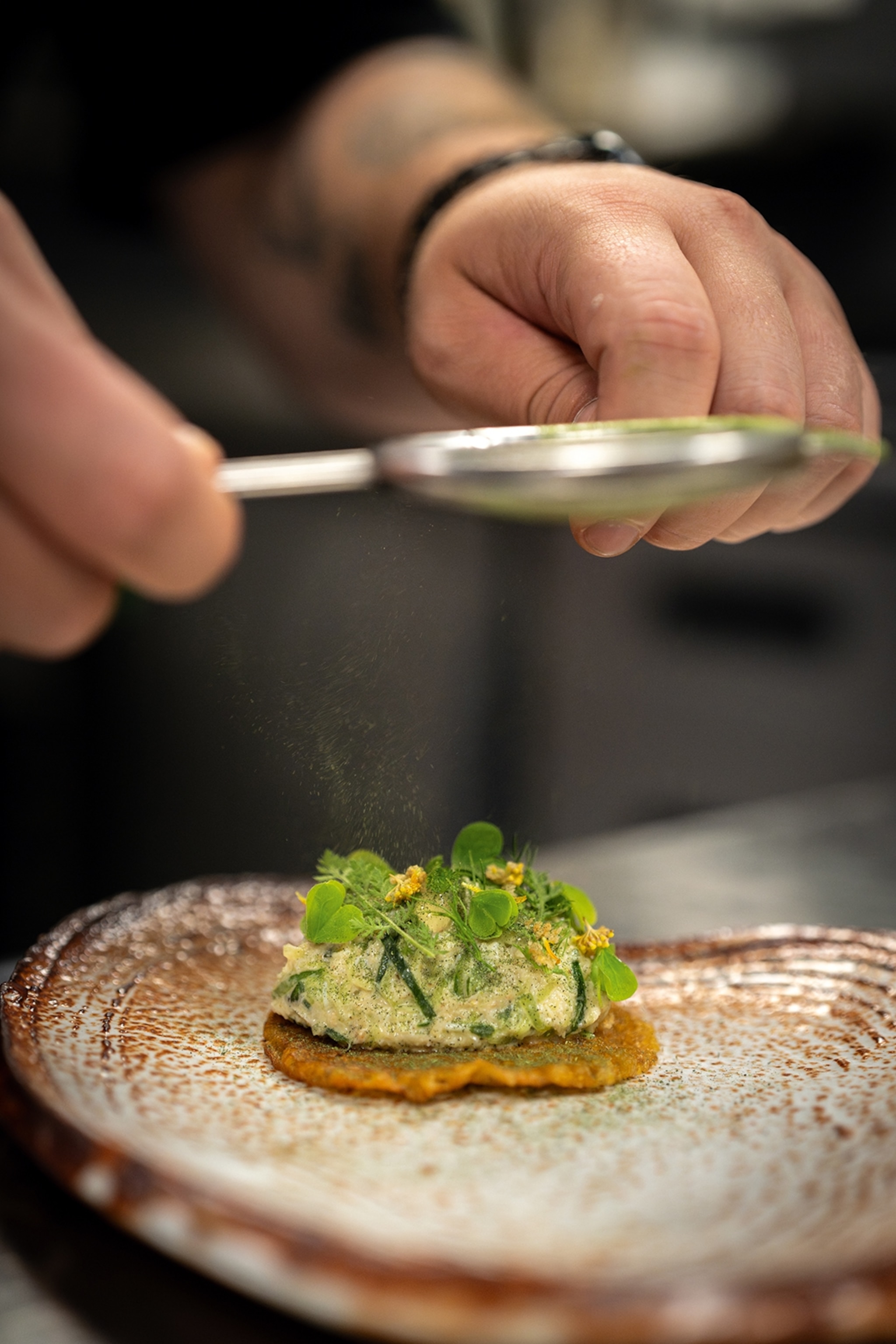 A close-up of a small lentil-based taco with a green filling and pea sprouts on top as a chef sieves matcha.