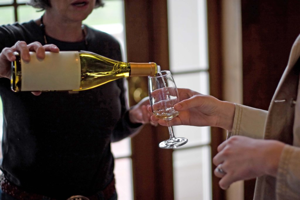 A woman pours white wine from a bottle into a tasting glass held by another person.