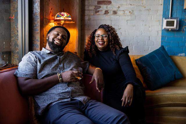 An intimate portrait of a smiling Black couple in a warmly lit lounge. The man, wearing a beanie, leans back with a drink in hand. The woman, with curly hair and glasses, sits beside him. They are on a comfortable couch, with a vintage Tiffany lamp casting a warm glow over the scene.