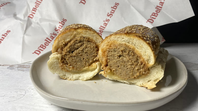 A DiBella's meatball sub with seeded bread split in half on a plate in front of the to-go packaging