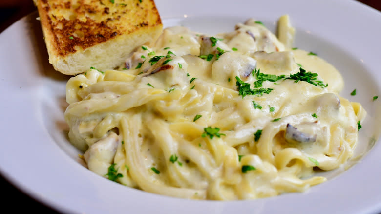White dish of fettuccine Alfredo pasta with parsley and a slice of garlic bread
